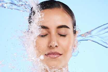 Water splash, face and skincare with a woman in studio on a blue background for hygiene or hydration. Relax, beauty and cleaning with an attractive young female in the shower for self care